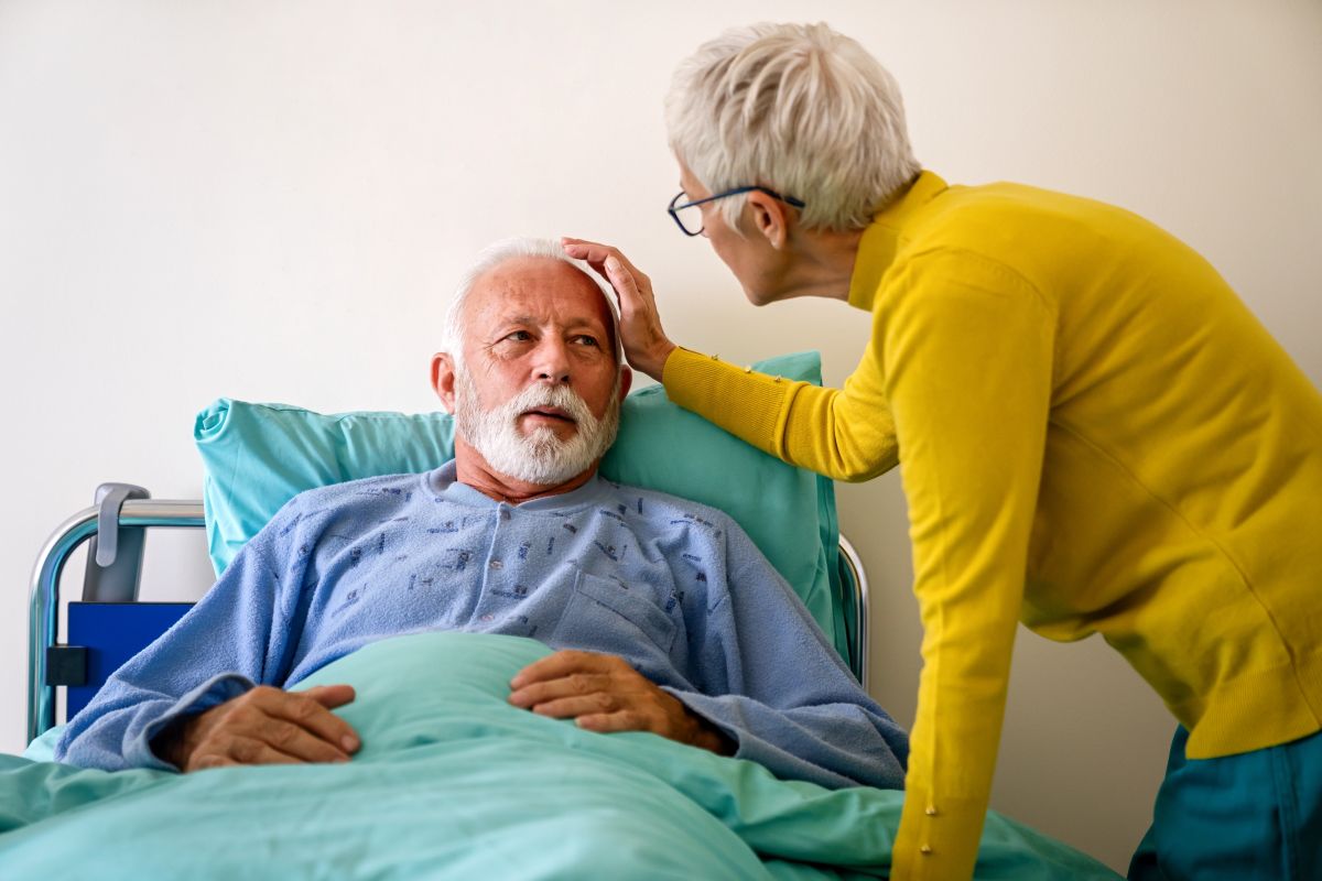 Older woman gently touching her seriously ill husband’s forehead as he rests in a hospital bed