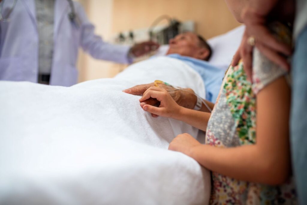 Family member resting a hand on the shoulder of a patient lying in a hospital bed with an IV line, with a clinician in the background